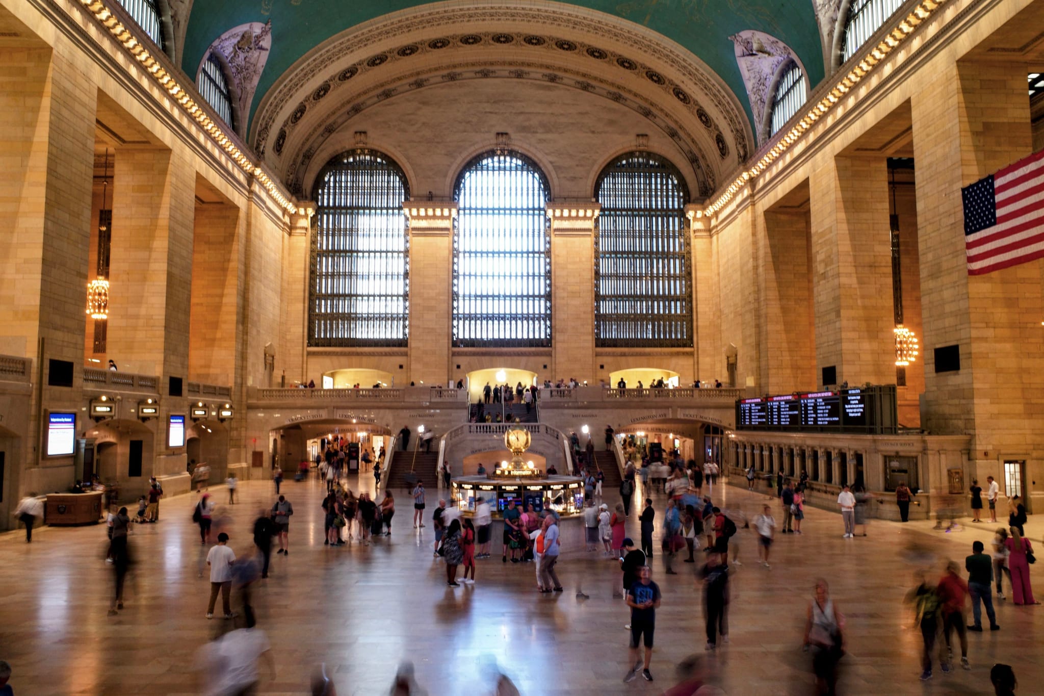 A bustling interior of a grand train station with high arched windows, a large American flag, and numerous people moving about