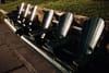 A row of dark green Adirondack chairs on a paved path beside grass, lit by low afternoon sun