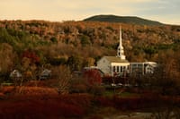A white church with a tall steeple nestled among autumn-colored trees at the foot of a hill, bathed in warm evening light
