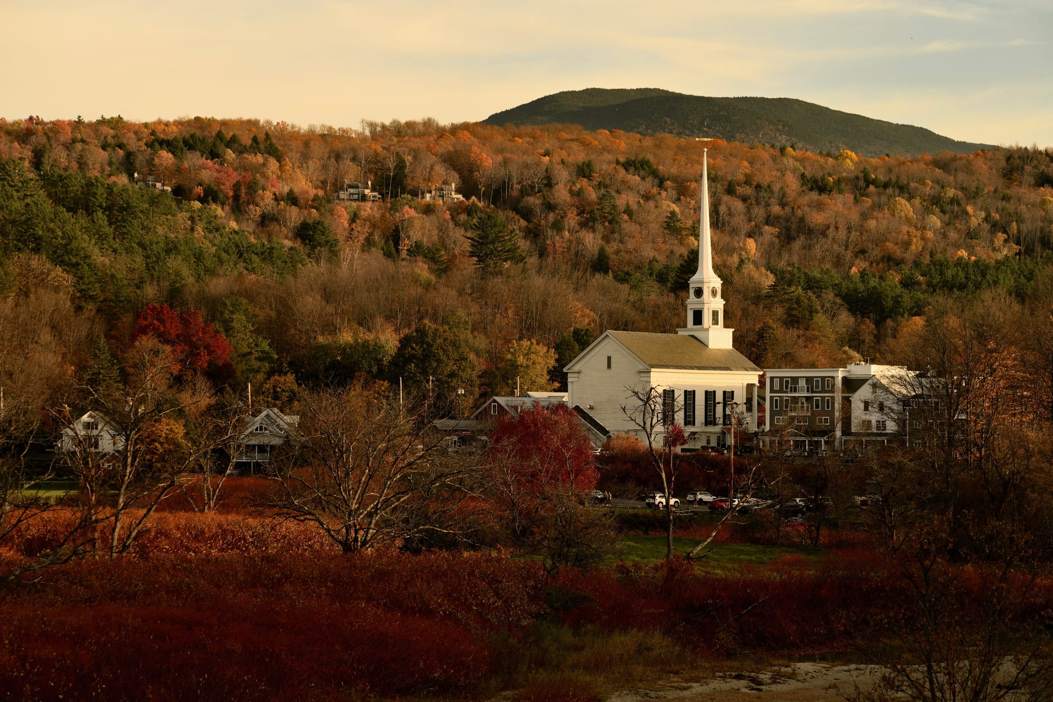 A white church with a tall steeple nestled among autumn-colored trees at the foot of a hill, bathed in warm evening light