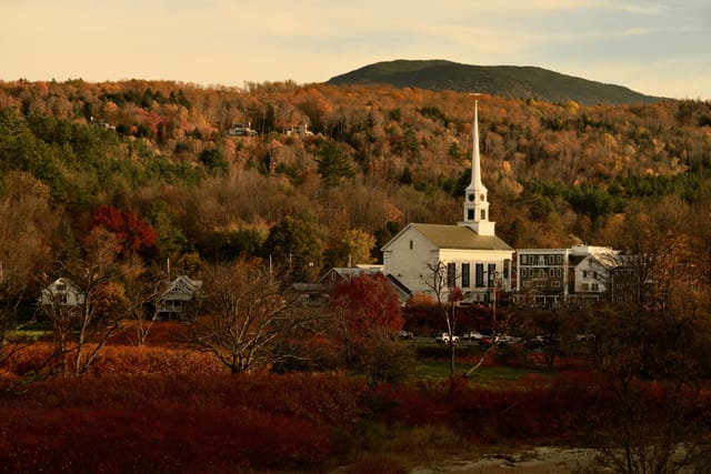 A white church with a tall steeple nestled among autumn-colored trees at the foot of a hill, bathed in warm evening light