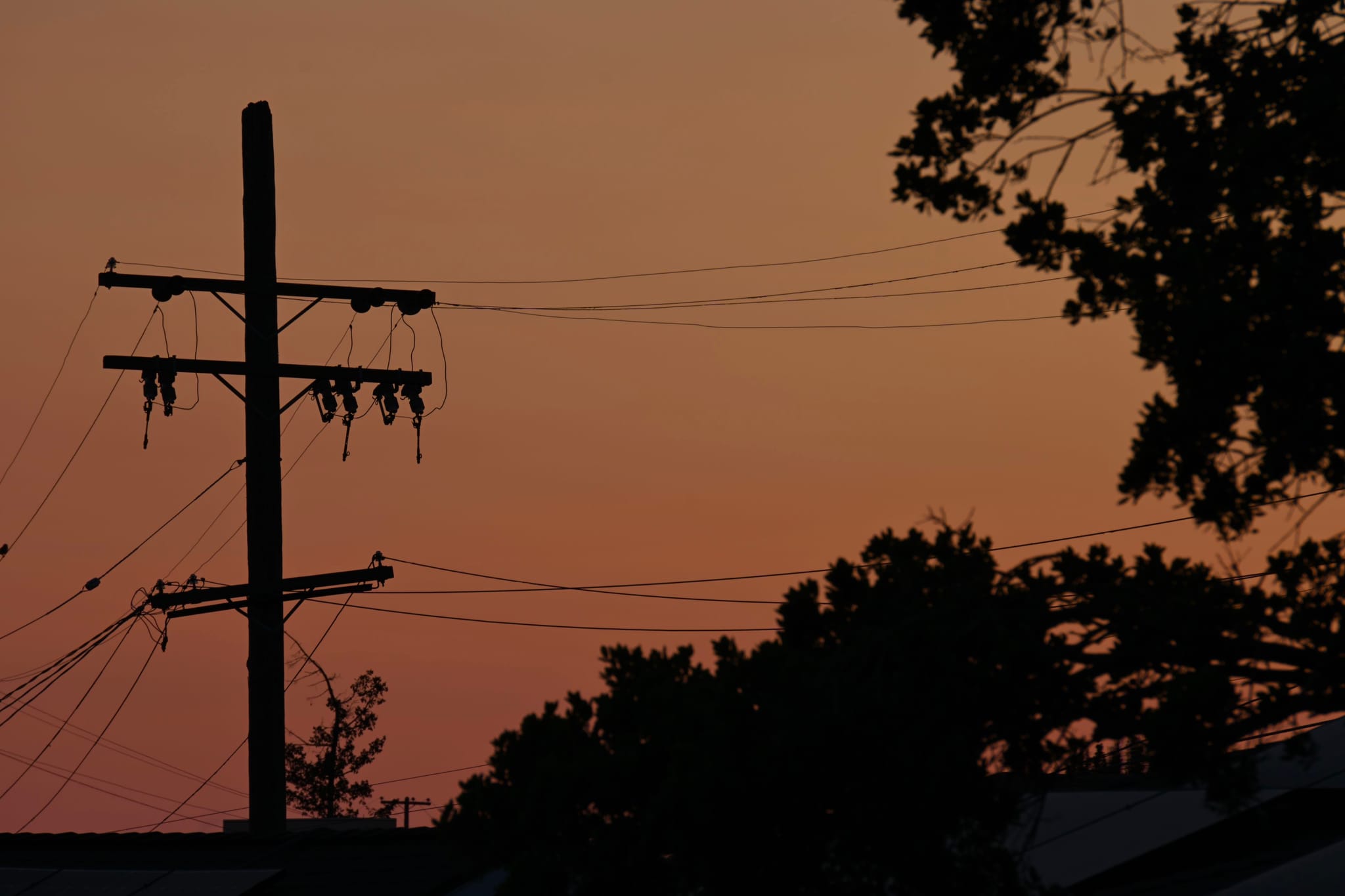 A silhouette of a utility pole and trees against a sunset sky with shades of orange and pink