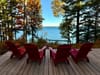 Four red Adirondack chairs on a wooden deck facing a calm lake, framed by tall trees with early autumn foliage