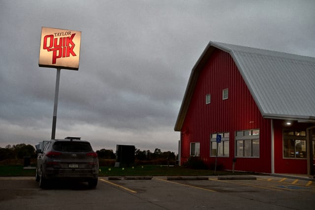 Red barn-style storefront with lit pole sign and a parked SUV in a nearly empty lot at dusk