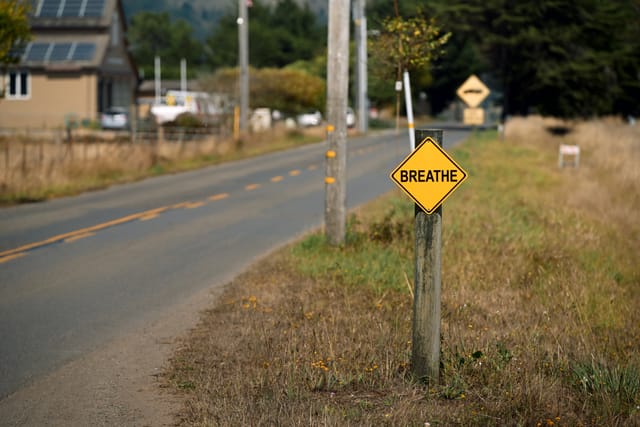 A rural road with a yellow diamond-shaped sign that reads BREATHE, surrounded by grass and trees, with houses in the background