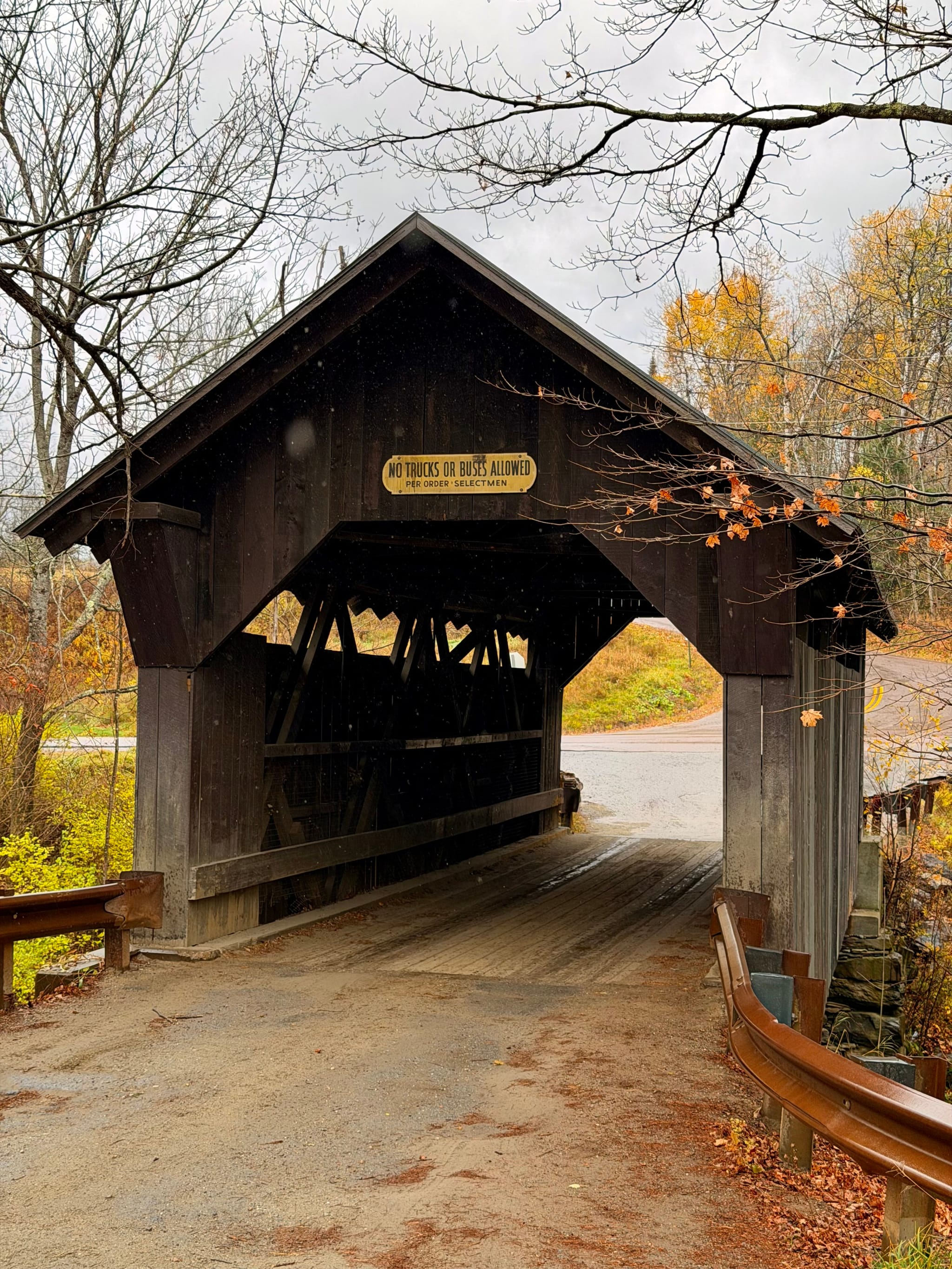 Rustic wooden covered bridge on a rainy autumn day, with wet road and colorful foliage in the background