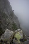 A climber ascends a steep, rocky mountain face shrouded in mist, with rugged terrain in the foreground
