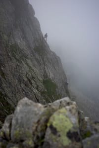 A climber ascends a steep, rocky mountain face shrouded in mist, with rugged terrain in the foreground