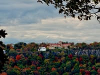 Large resort perched on a cliff above colorful autumn forest, framed by overhanging branches under a cloudy sky