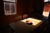 A dimly lit room with a wooden table and chairs, featuring a bowl on the table. Sunlight streams through a window, casting shadows across the table