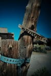 A weathered wooden post with a metal band and a broken sign, set against a clear blue sky and rustic buildings in the background