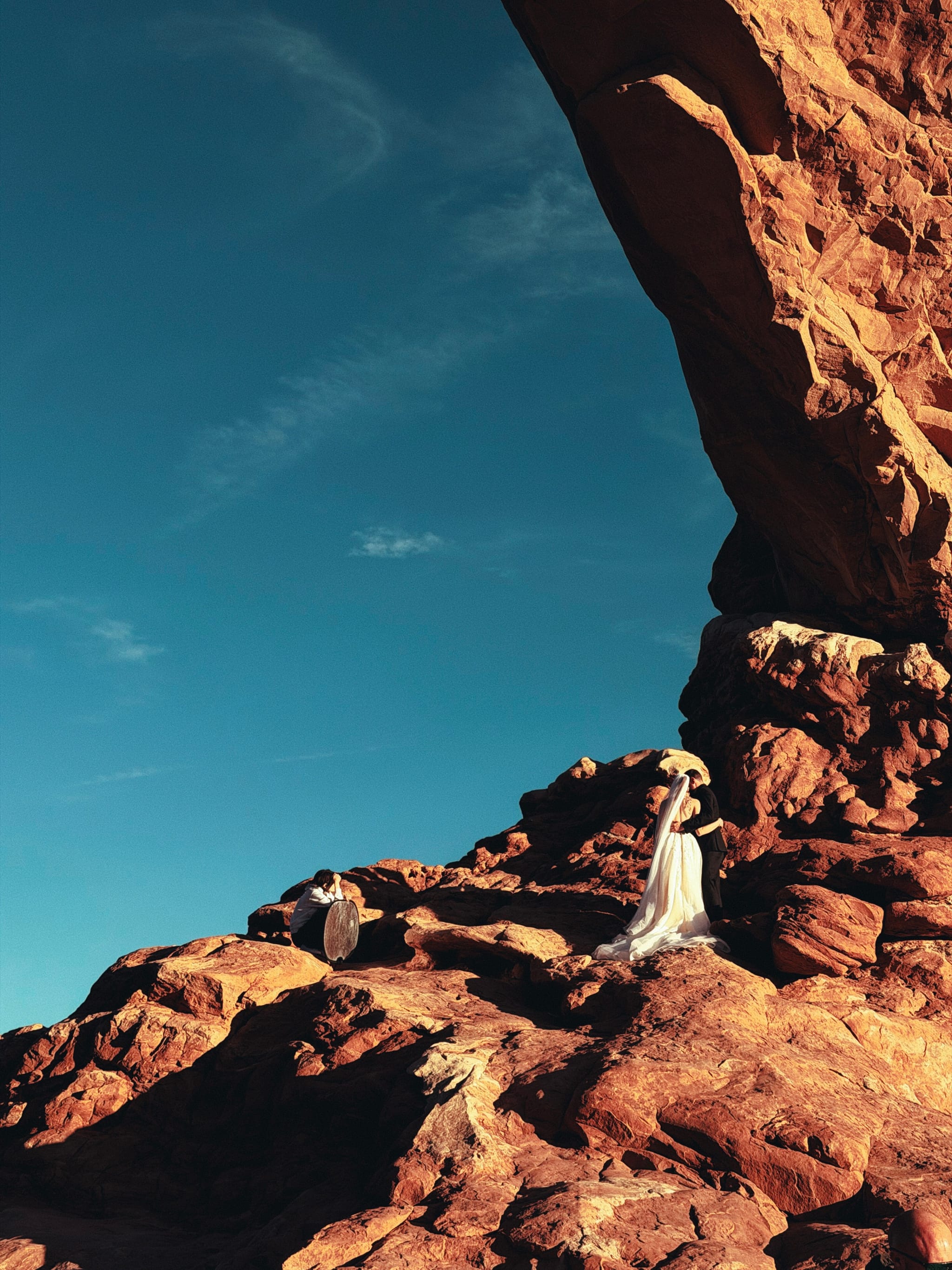 Person in white dress standing on sunlit sandstone beneath a towering rock arch under clear blue sky