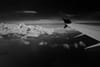 A black and white view from an airplane window, featuring the aircraft's wing and a vast expanse of clouds below