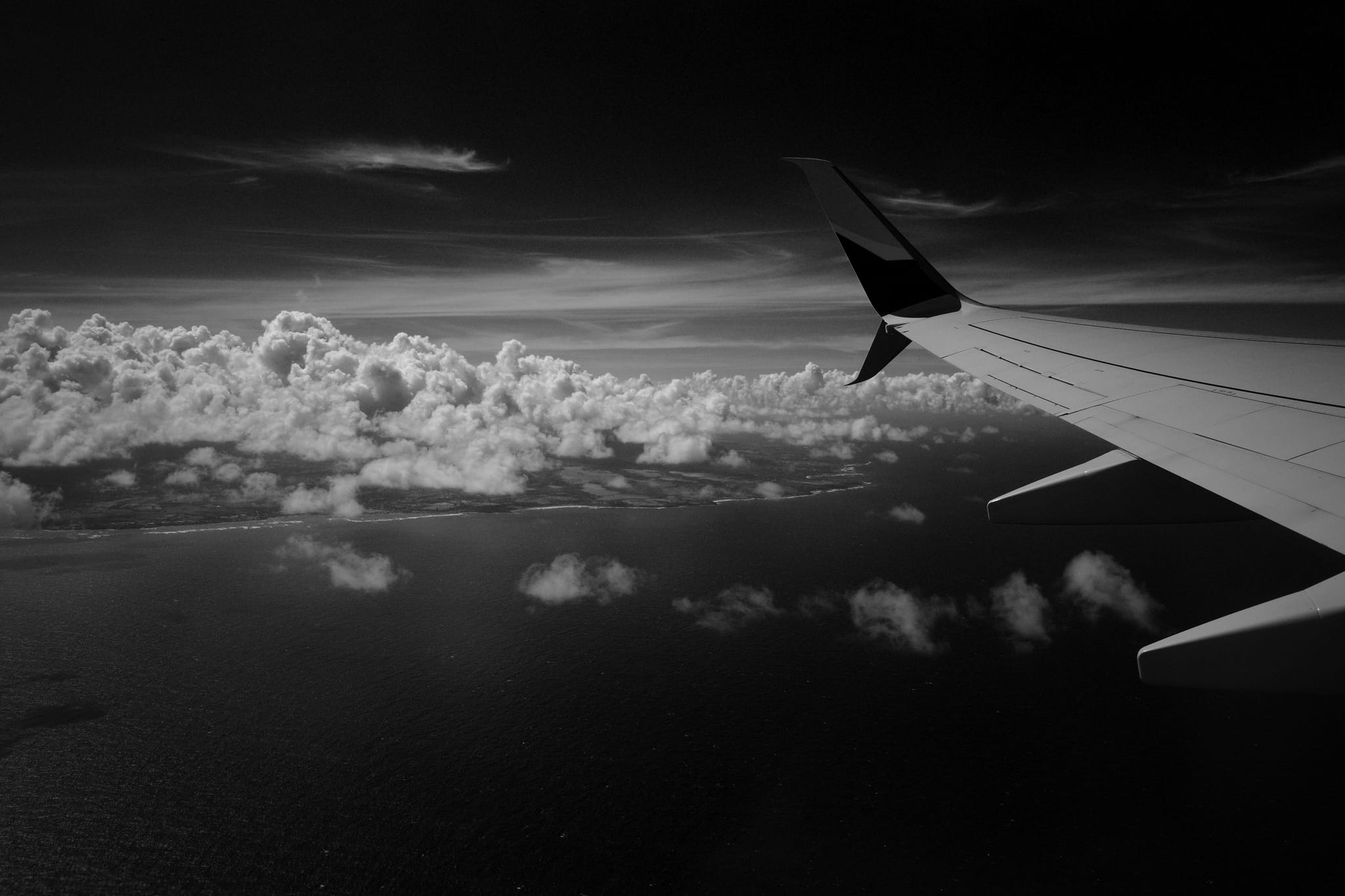 A black and white view from an airplane window, featuring the aircraft's wing and a vast expanse of clouds below