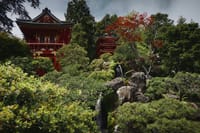 A traditional Japanese garden with lush greenery, a small waterfall, and a red pagoda-style building in the background