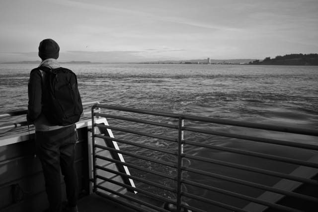 A person with a backpack stands on a ferry, gazing out at the water and distant shoreline under a cloudy sky. The scene is in black and white