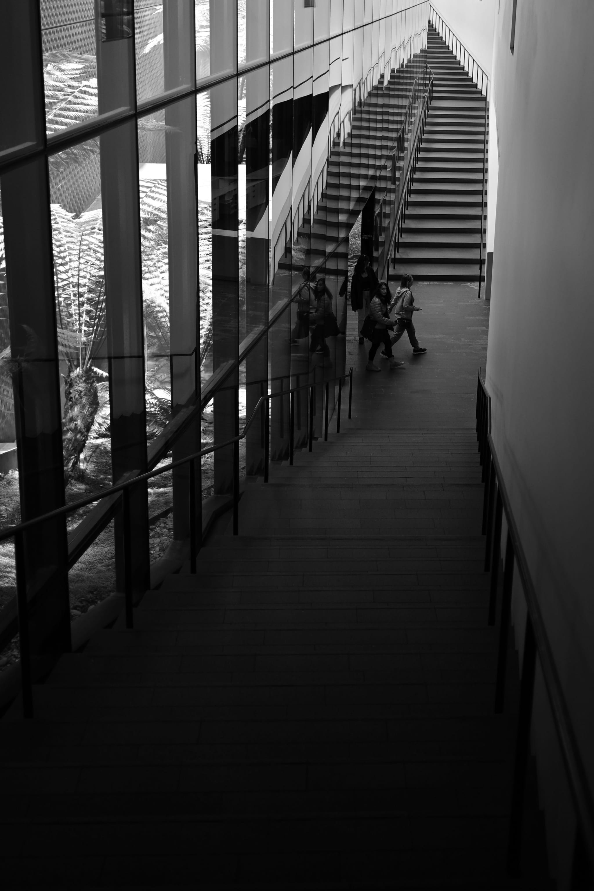 A black and white photograph of a modern staircase with glass railings, featuring a few people ascending. The perspective creates a dramatic, angular composition with reflections on the glass