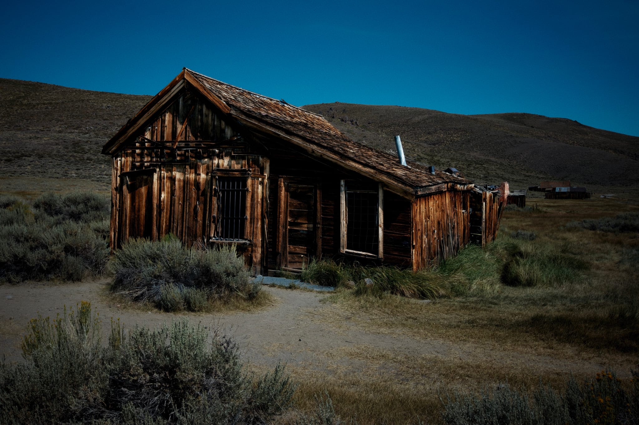 An old, weathered wooden cabin stands in a desolate, grassy landscape under a clear blue sky