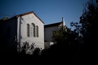 A white house with arched windows is partially illuminated by sunlight, surrounded by dark foliage and set against a clear blue sky