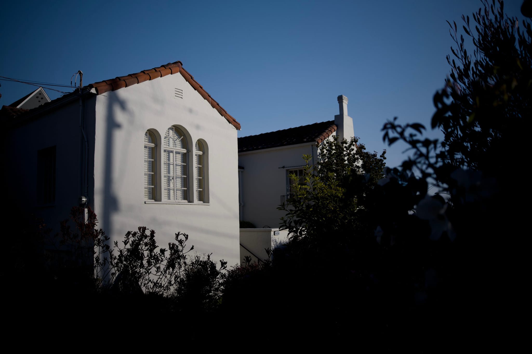 A white house with arched windows is partially illuminated by sunlight, surrounded by dark foliage and set against a clear blue sky