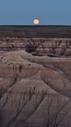 Full moon above a rugged, layered badlands landscape at dusk