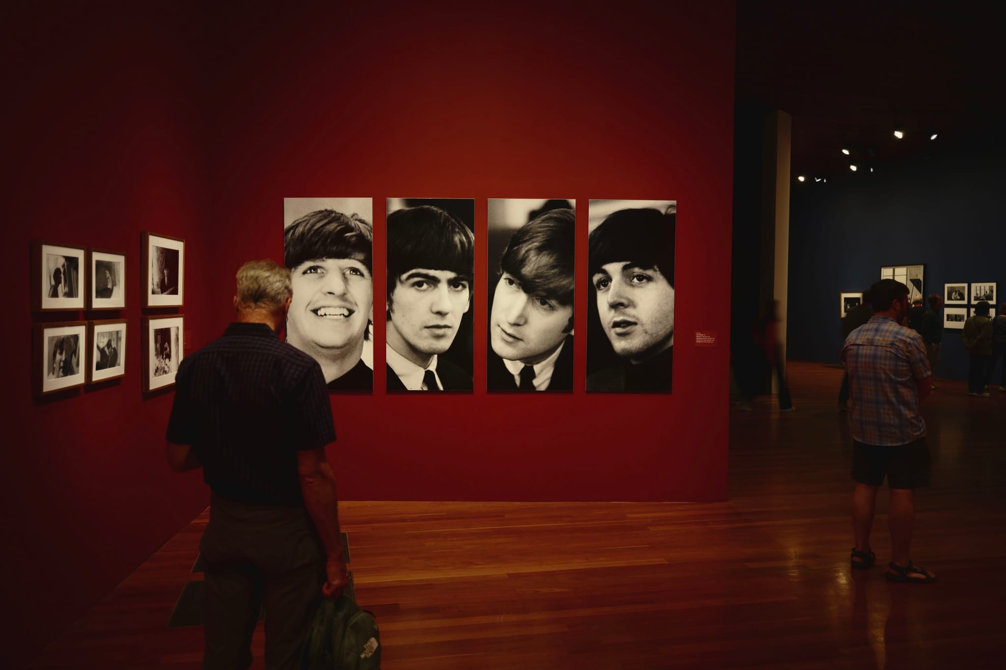 A gallery with large black-and-white portraits of four individuals on a red wall, with visitors observing the artwork