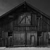 A black and white photograph of a rustic wooden barn with a gabled roof, featuring large double doors and several small windows