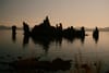 Silhouetted rock formations rise from a calm body of water at dusk, with distant mountains and a soft, dimly lit sky in the background