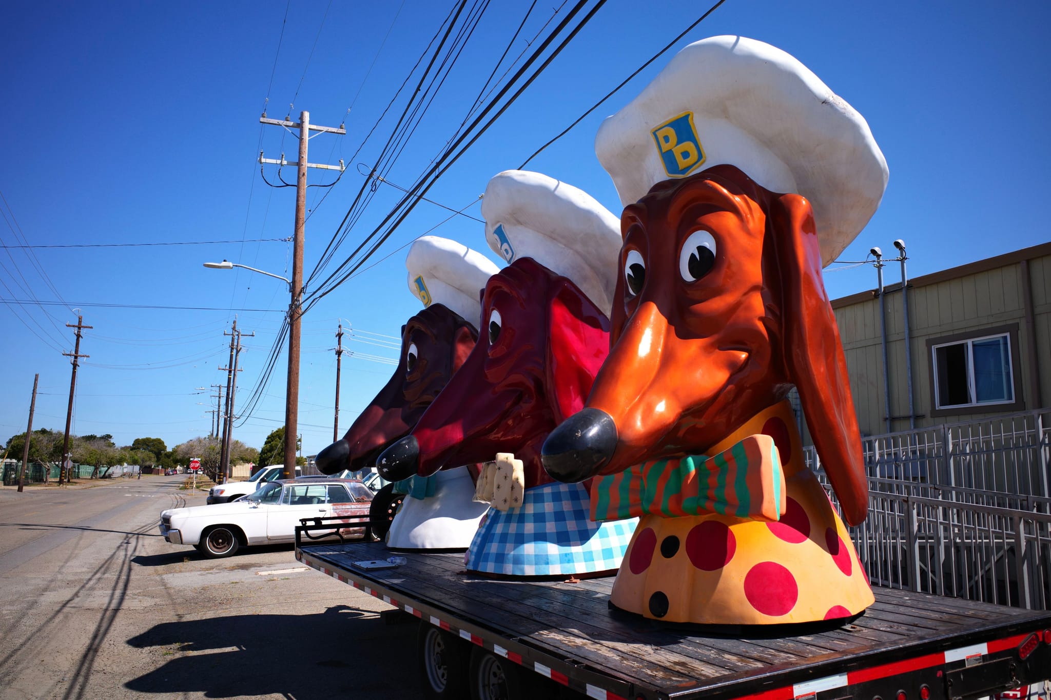 Large dachshund statues wearing chef hats and scarves are displayed on a flatbed trailer in an industrial area under a clear blue sky