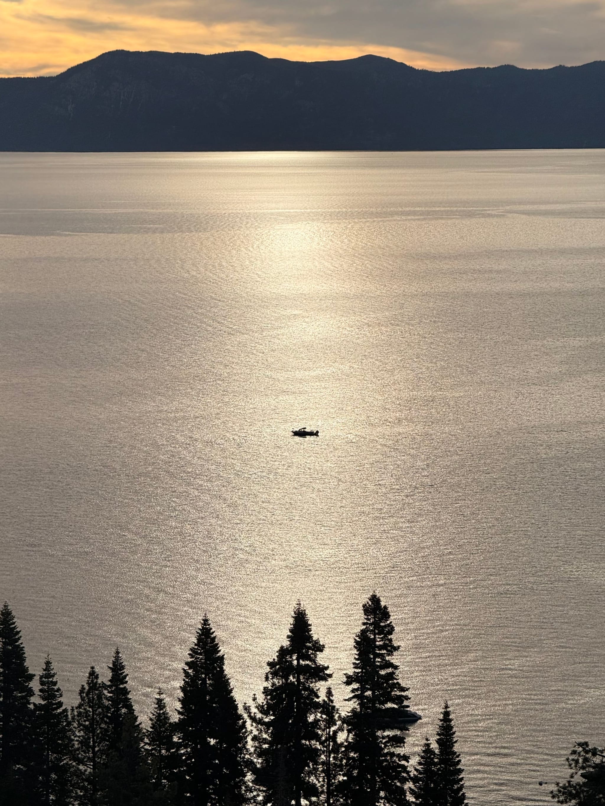 A serene lake at sunset with a small boat in the center, surrounded by silhouetted trees and distant mountains