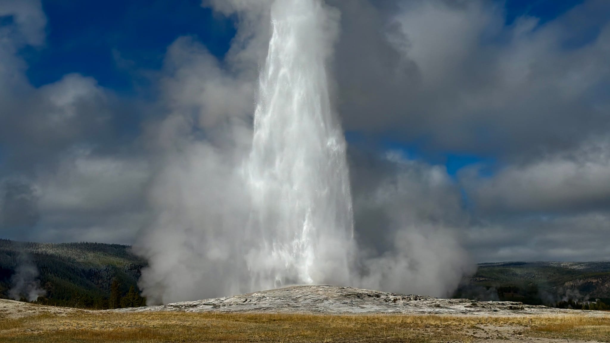 A powerful geyser erupts, sending a tall plume of water and steam into a cloudy sky over a rocky basin