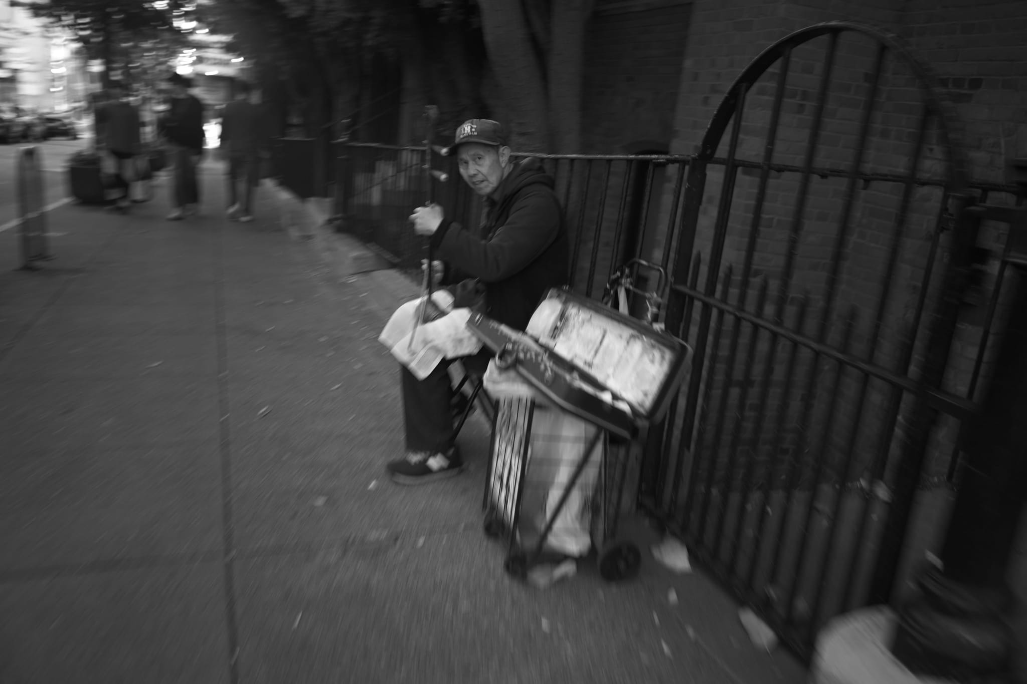 A person sitting on a sidewalk near a fence, holding a newspaper and a drink, with a cart beside them. The image is in black and white and slightly blurred