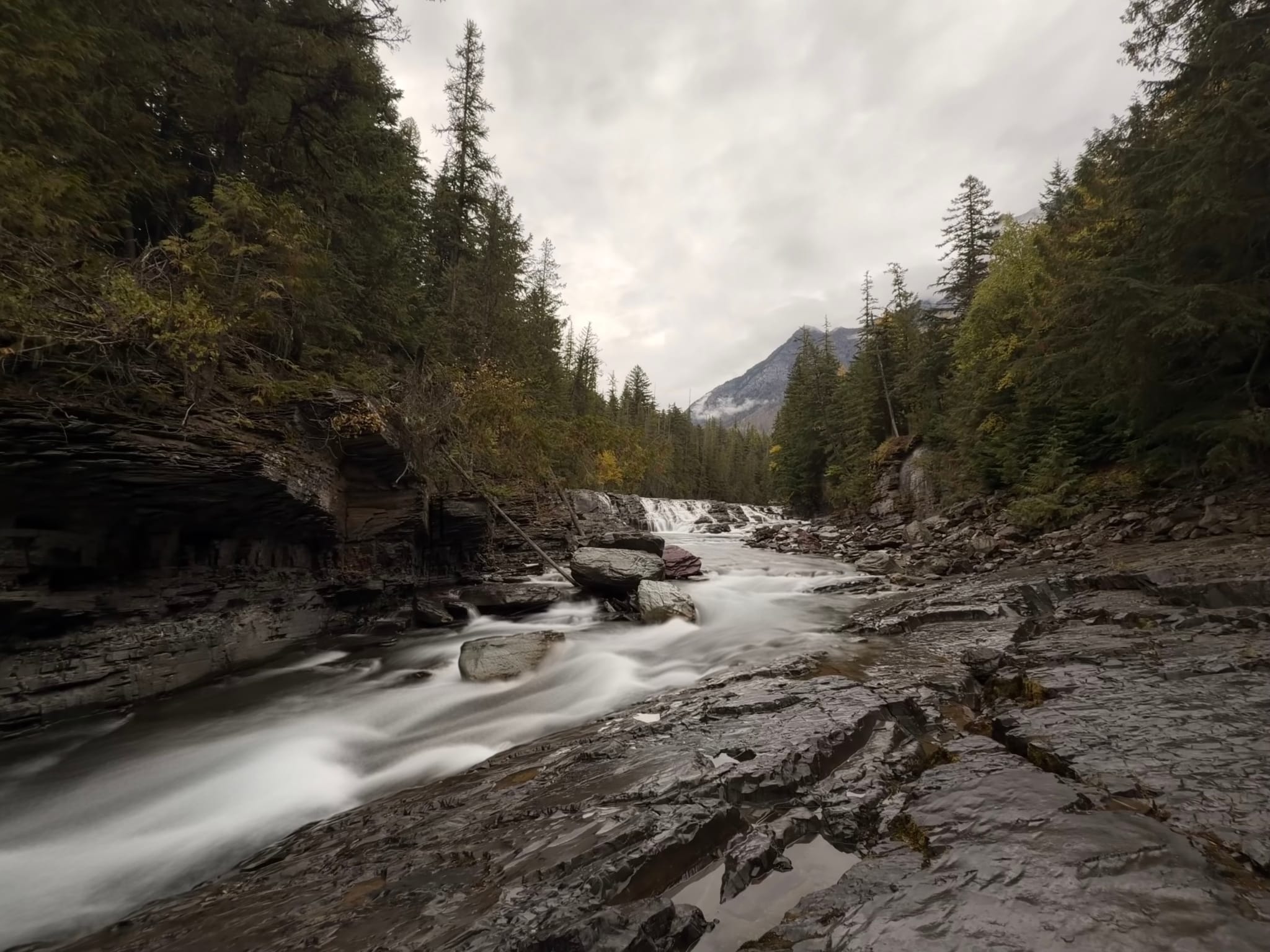 Long-exposure view of a rushing river over slick rock slabs in a forested valley, with a distant mountain beneath an overcast sky