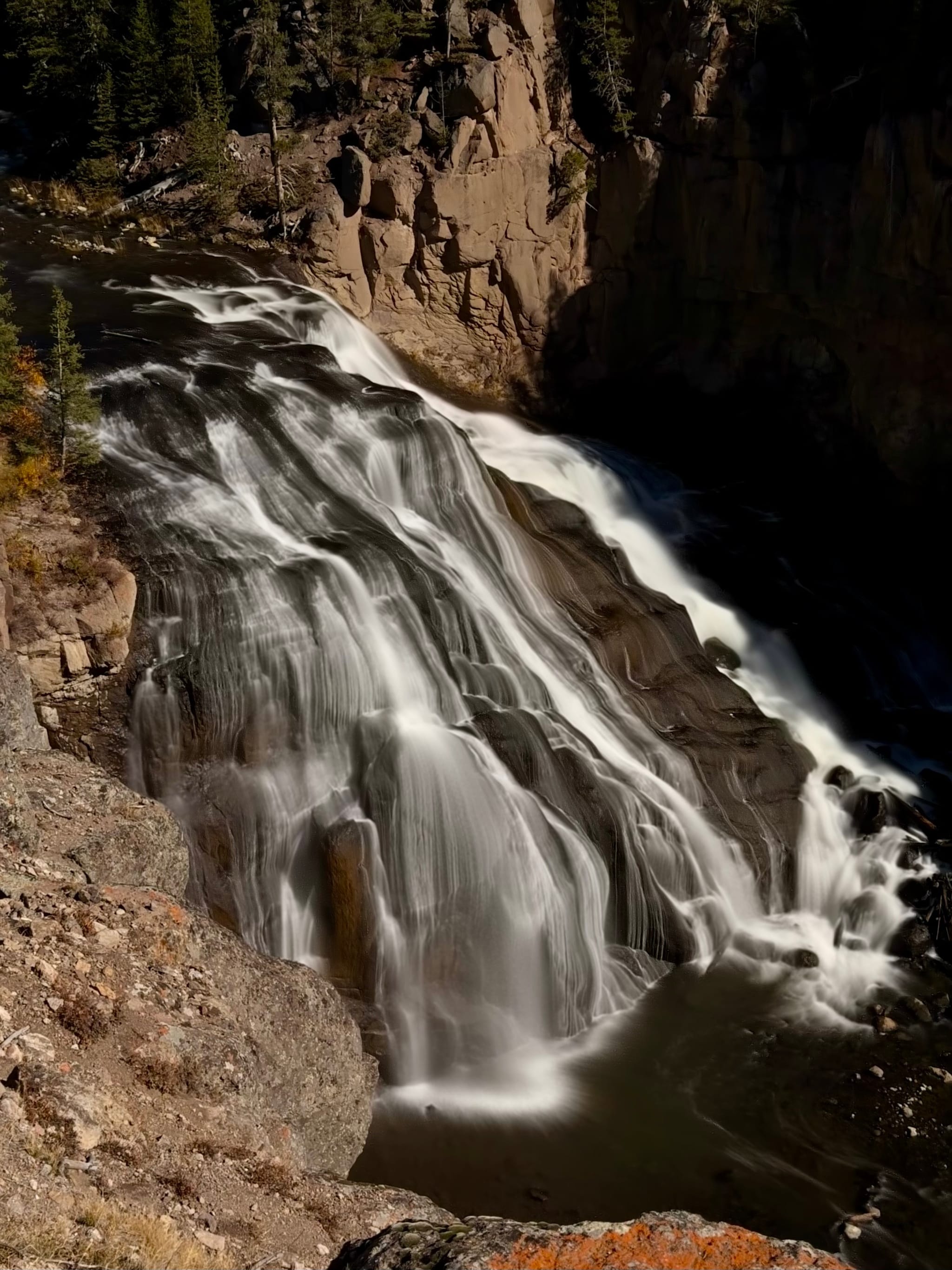 A tiered waterfall cascading over dark rock in a narrow canyon, captured with a long exposure, with pine trees along the rim