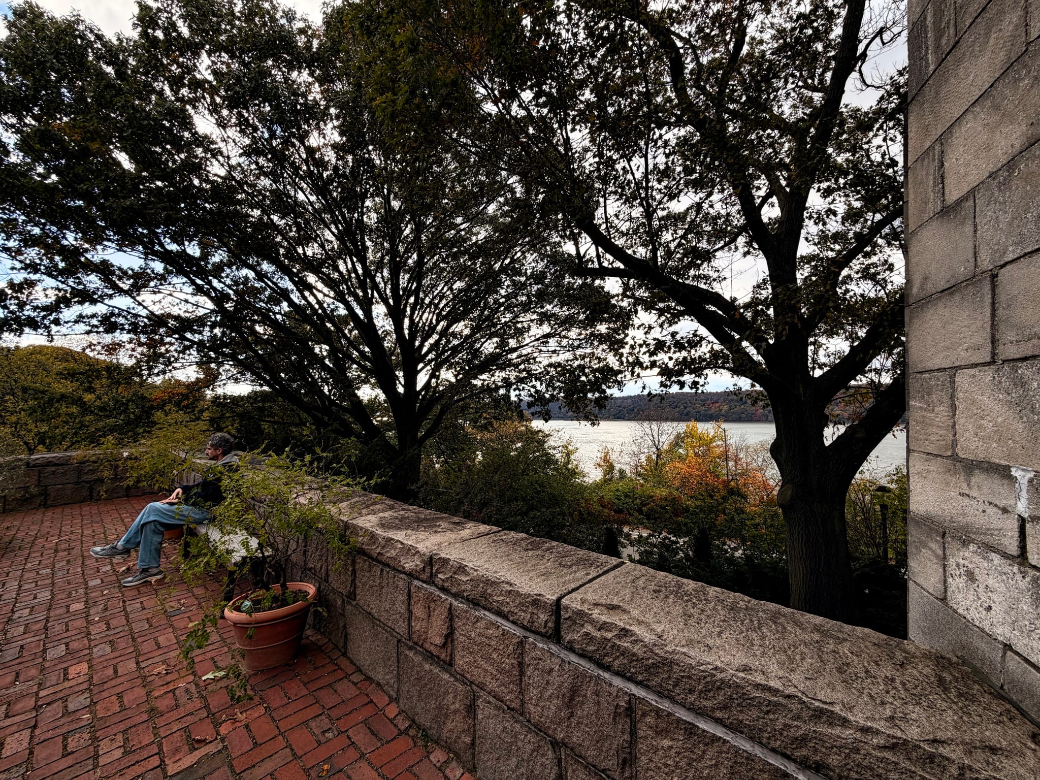 Brick patio beside stone wall with potted plant and a seated person, trees framing a river view