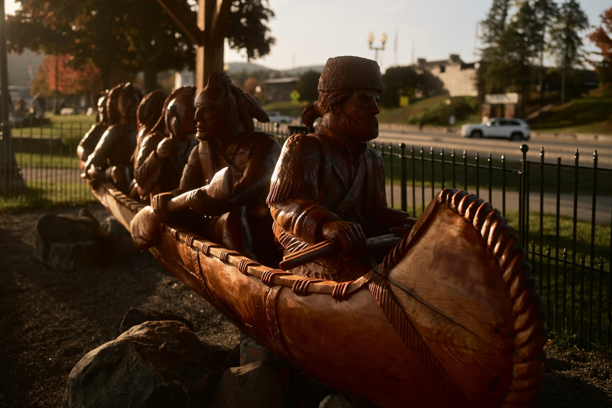 Wooden sculpture of several figures paddling a canoe, set outdoors near a roadside fence in warm late-day light