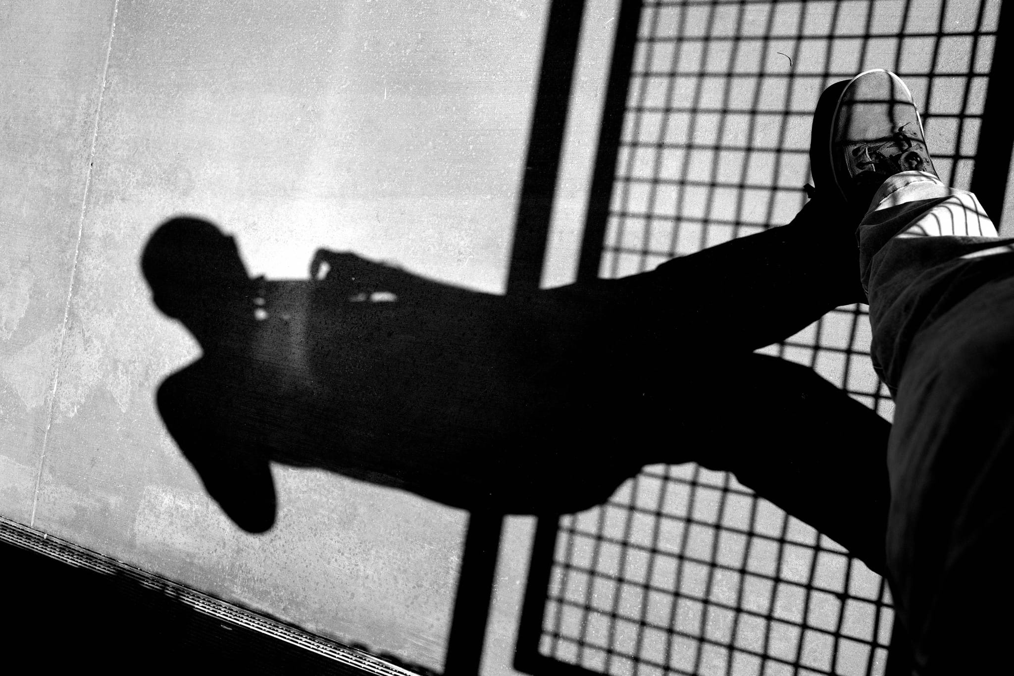 A black and white reflection of a person taking a photo, with their shadow cast on a wet surface next to a grid-like structure