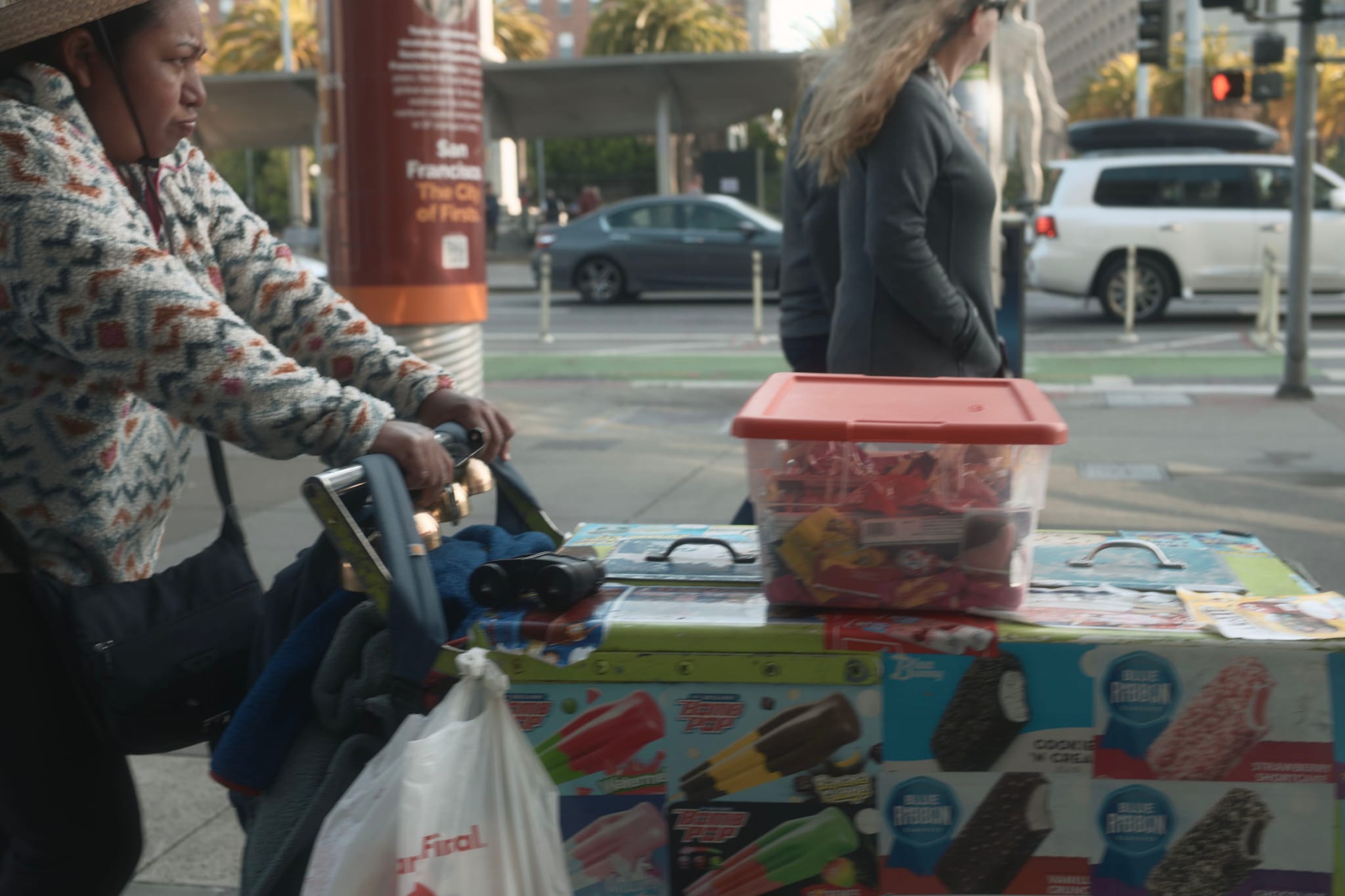 A street vendor pushes a cart covered with images of ice cream, carrying a red-lidded container and a bag, while a person walks by on the sidewalk