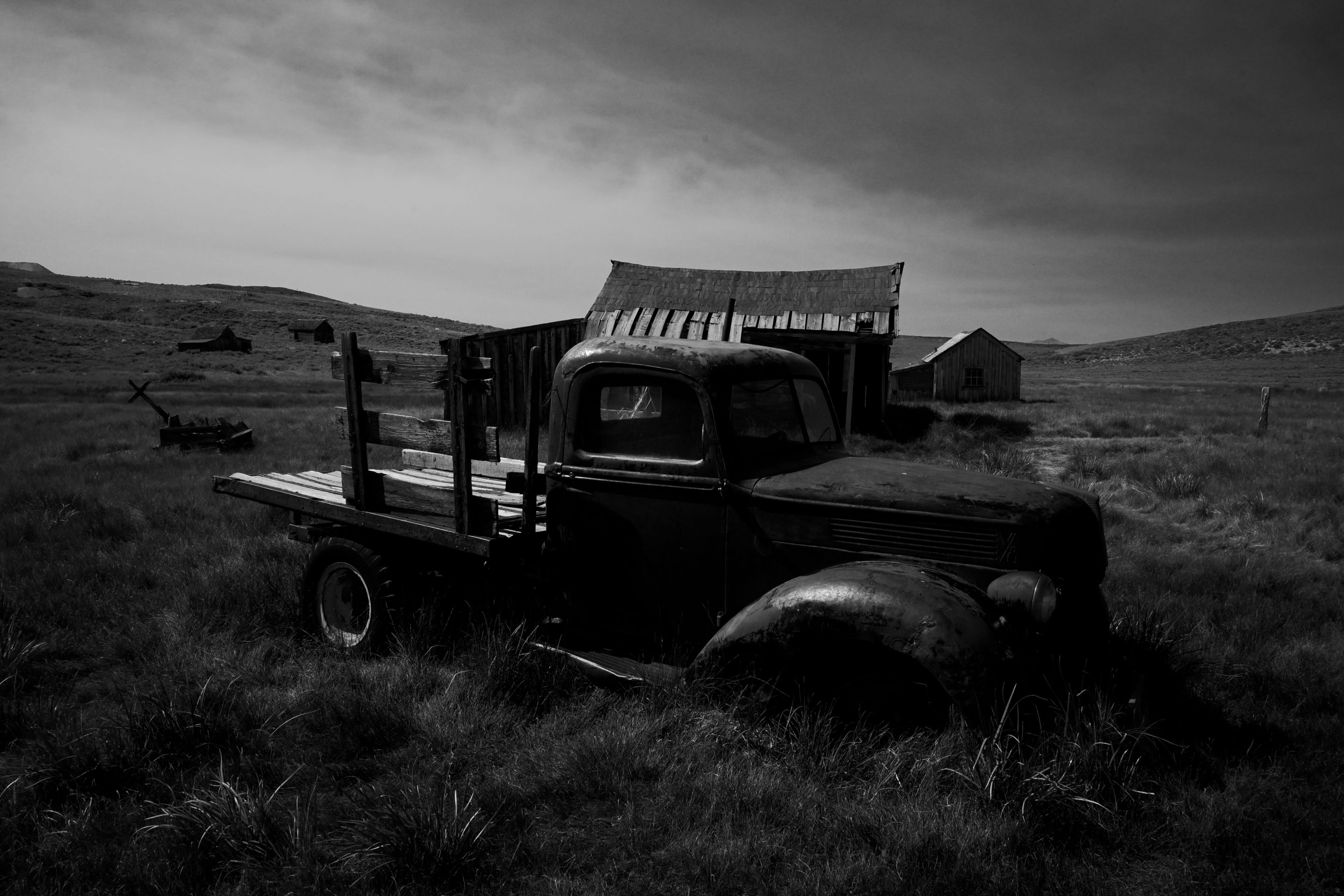 A black and white scene featuring an old, abandoned truck in a grassy field with a rustic barn and distant hills under a cloudy sky