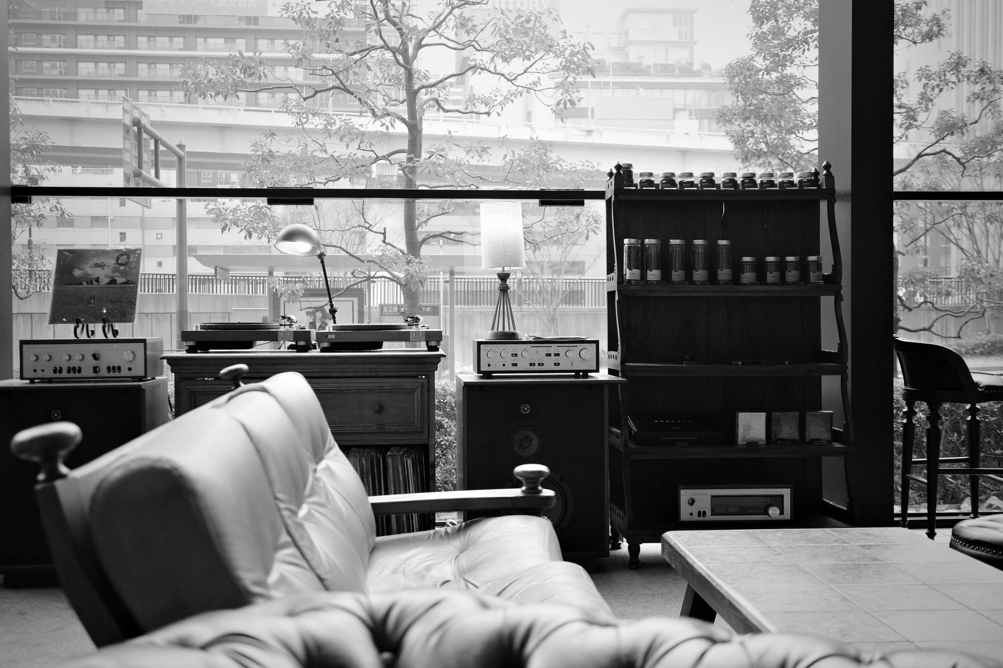 A black and white interior scene with a leather sofa, a wooden table, and a shelf with various items. Large windows reveal an urban view with trees and buildings outside