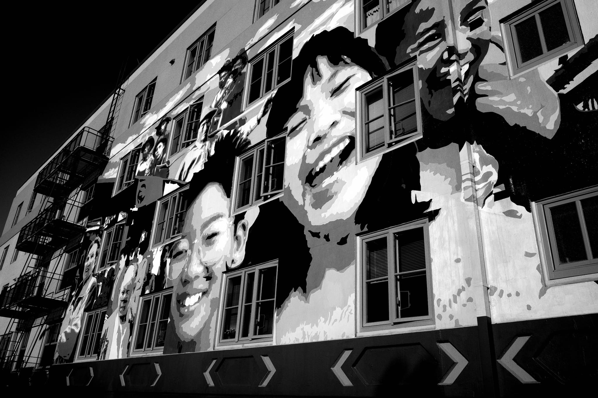A black and white mural on a building features large, expressive faces of smiling individuals, with windows integrated into the artwork