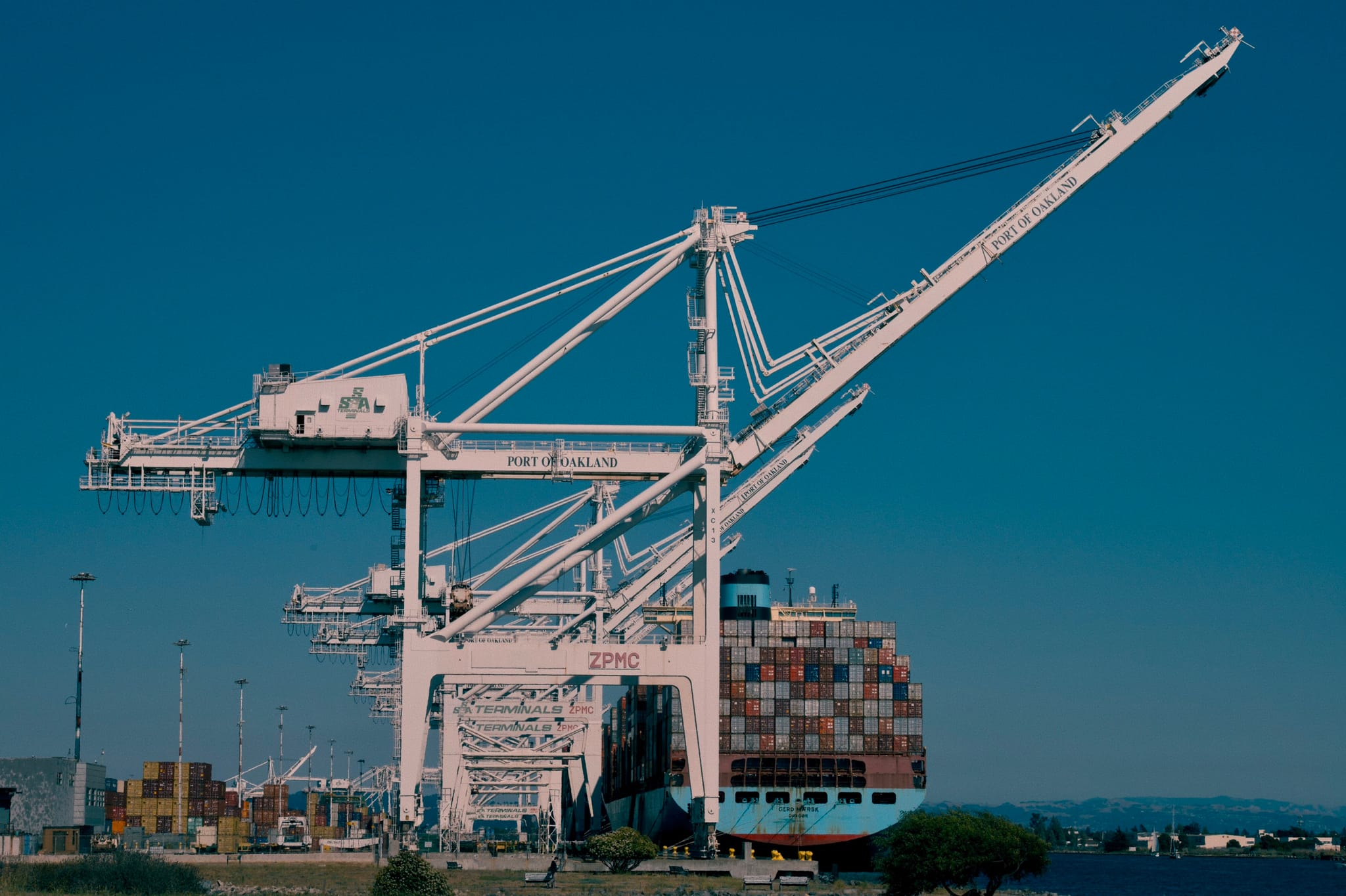 A large crane at a port, with a container ship docked nearby under a clear blue sky