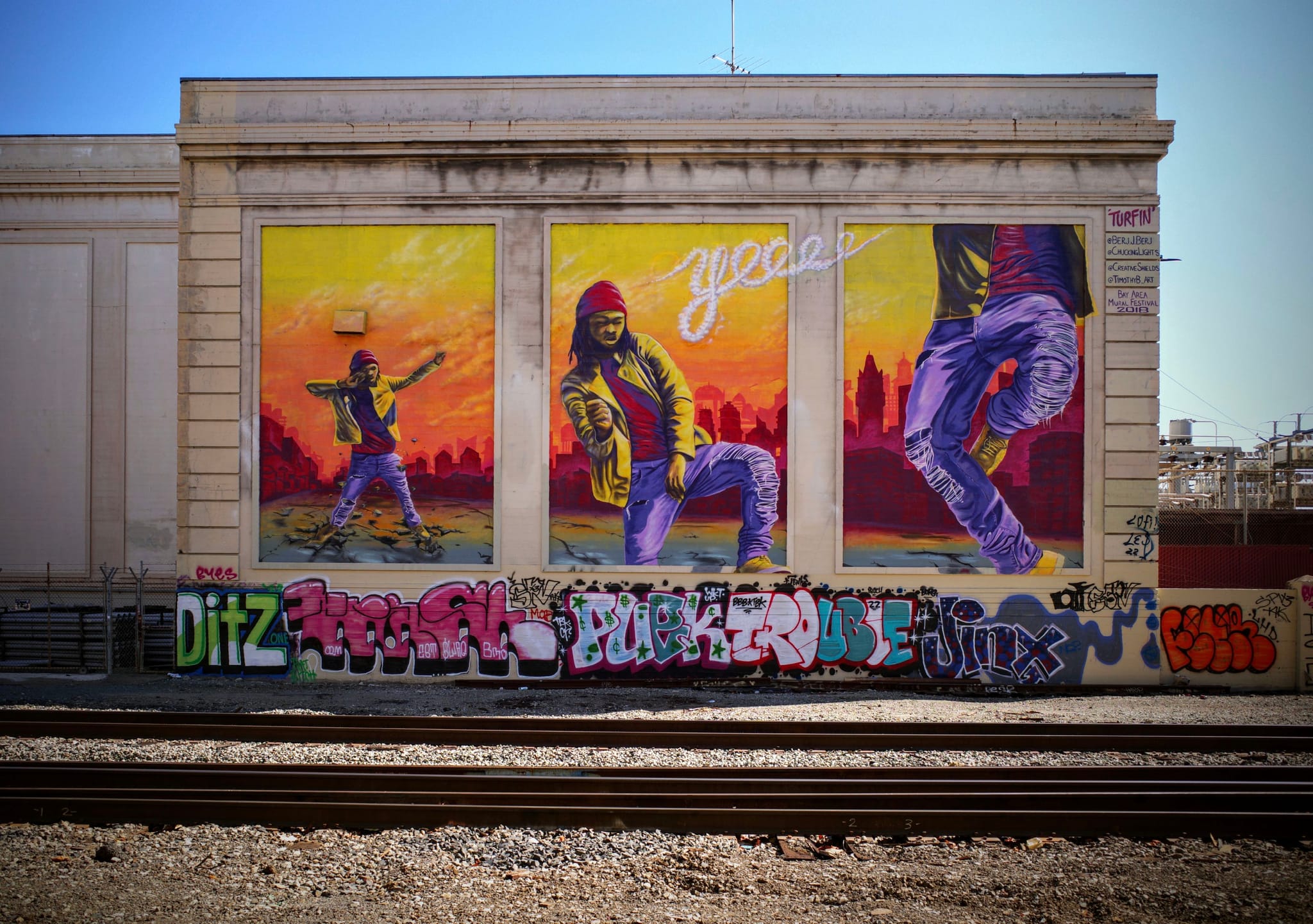A vibrant mural on a building wall features three panels depicting dancers in dynamic poses against a colorful background. The word groove is prominently displayed above the central figure. Below the mural, a row of graffiti covers a low wall near train tracks