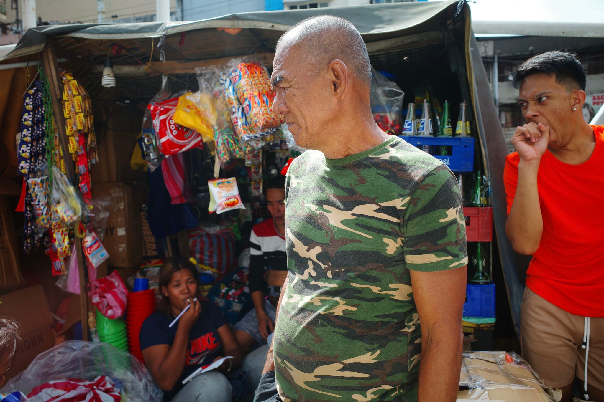 A bustling market scene with people interacting near a small stall displaying various goods. One person in a camouflage shirt stands in the foreground, while others are seated or standing nearby, surrounded by colorful items