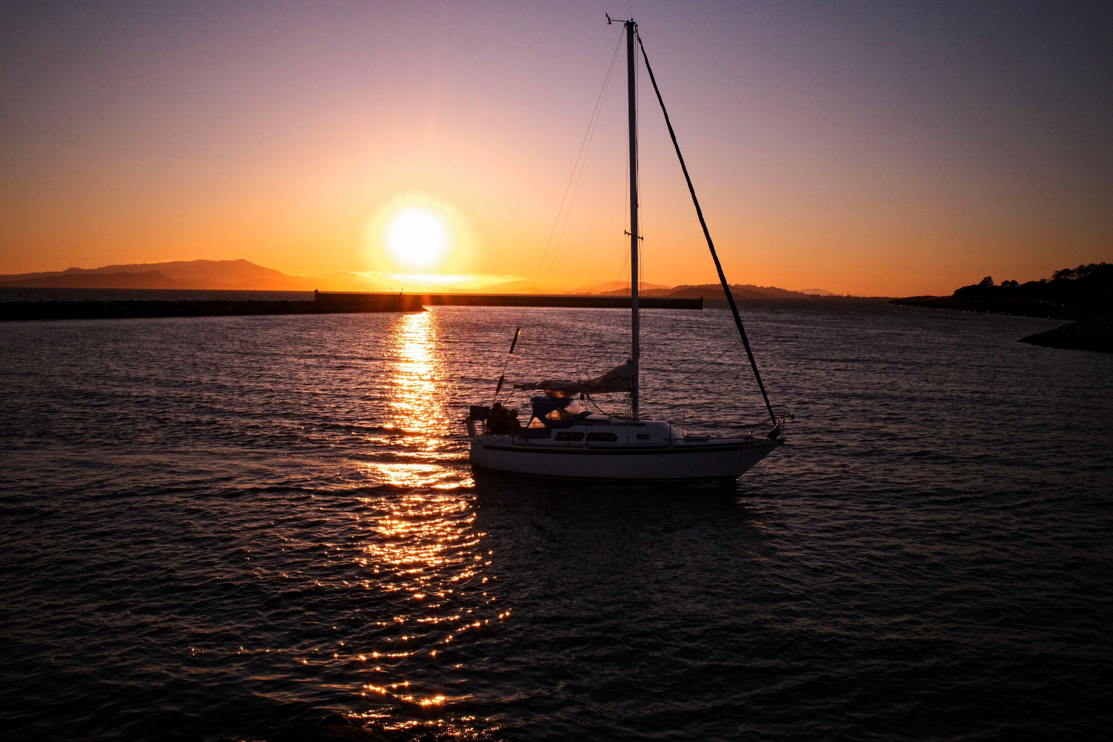 A sailboat on calm water during a vibrant sunset, with the sun low on the horizon and a silhouette of distant hills