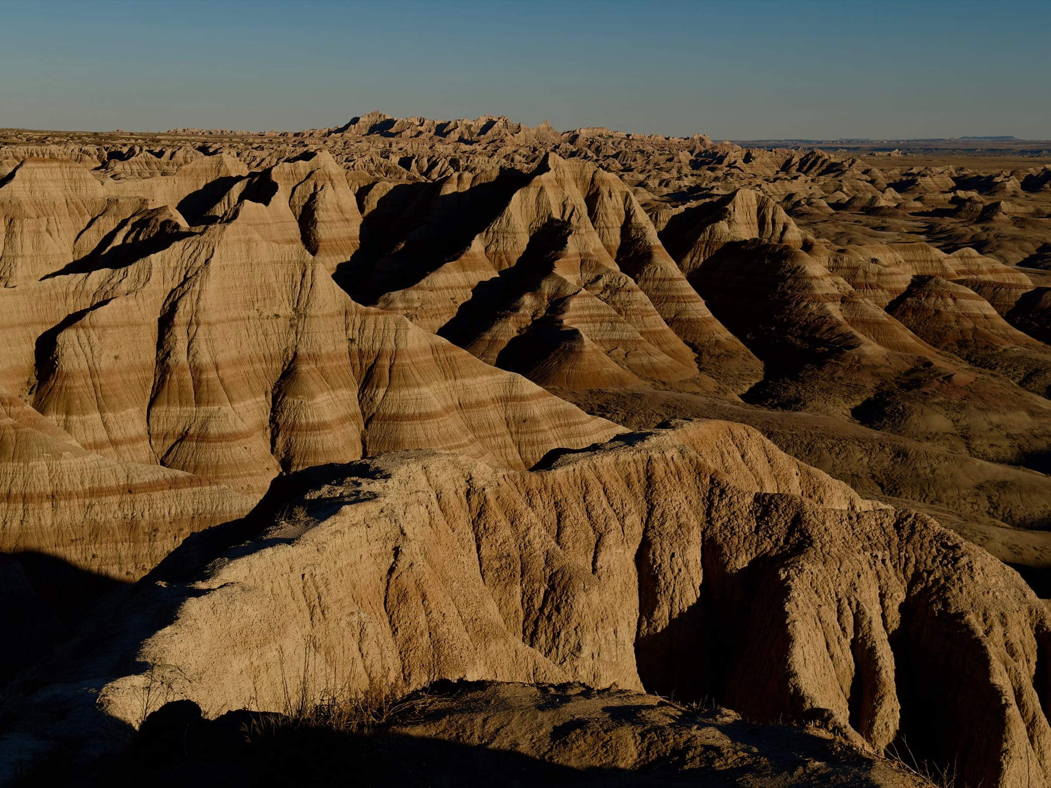 Rugged badlands with layered rock ridges and gullies, lit by low sun casting long shadows across the arid terrain