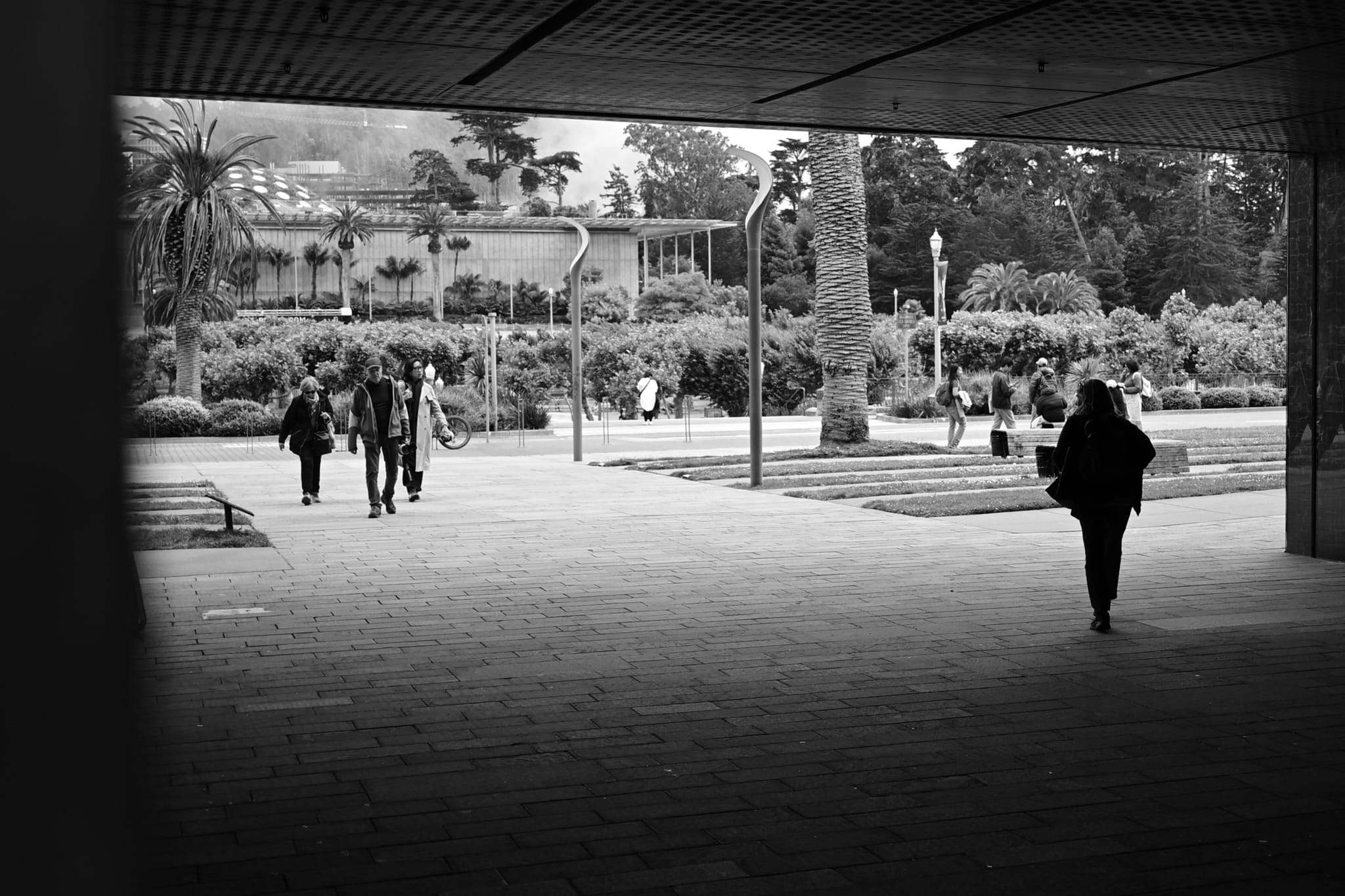 A black and white scene of people walking in an outdoor area with a building and trees in the background, partially framed by a dark overhang