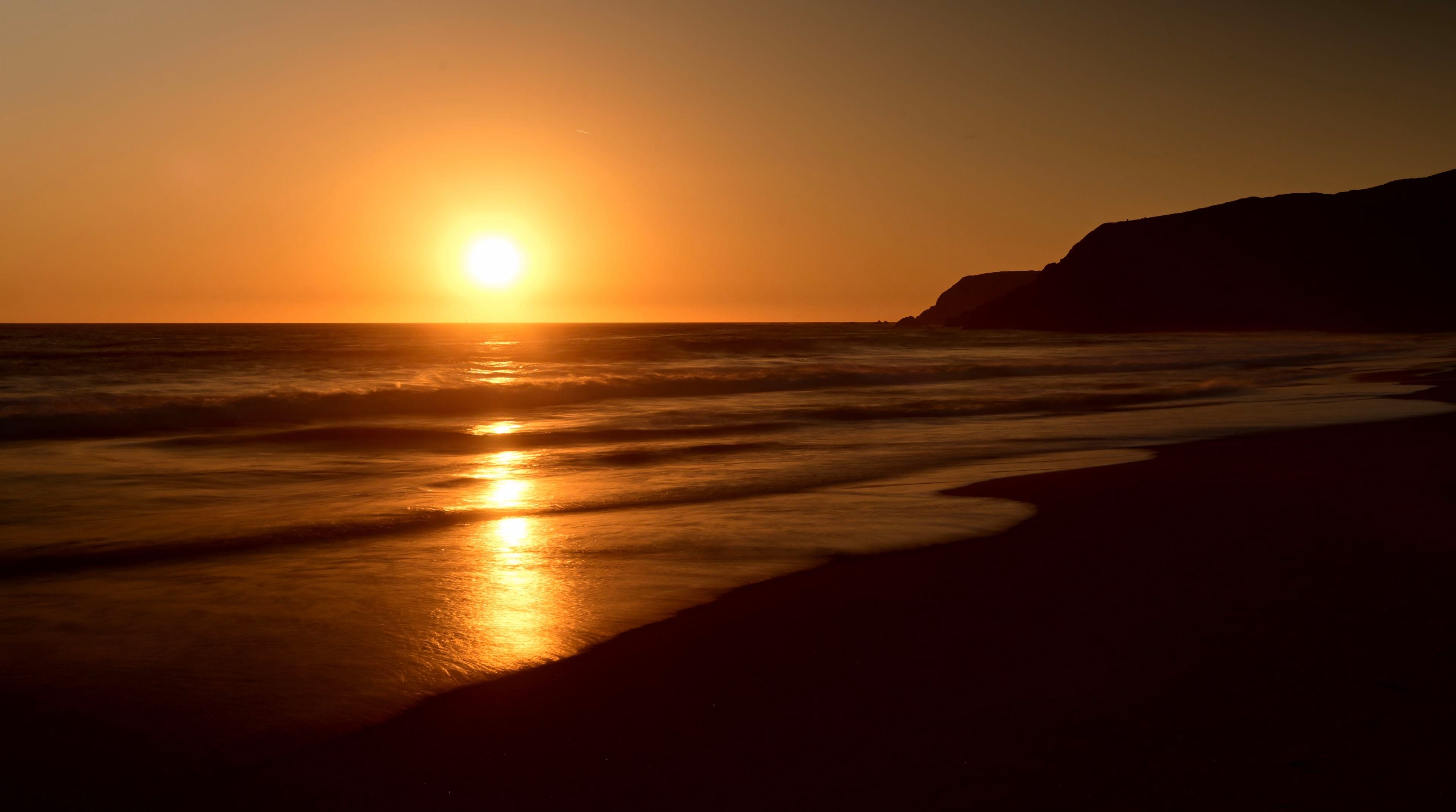A serene sunset over the ocean with gentle waves lapping against a sandy beach and a silhouette of a distant hill