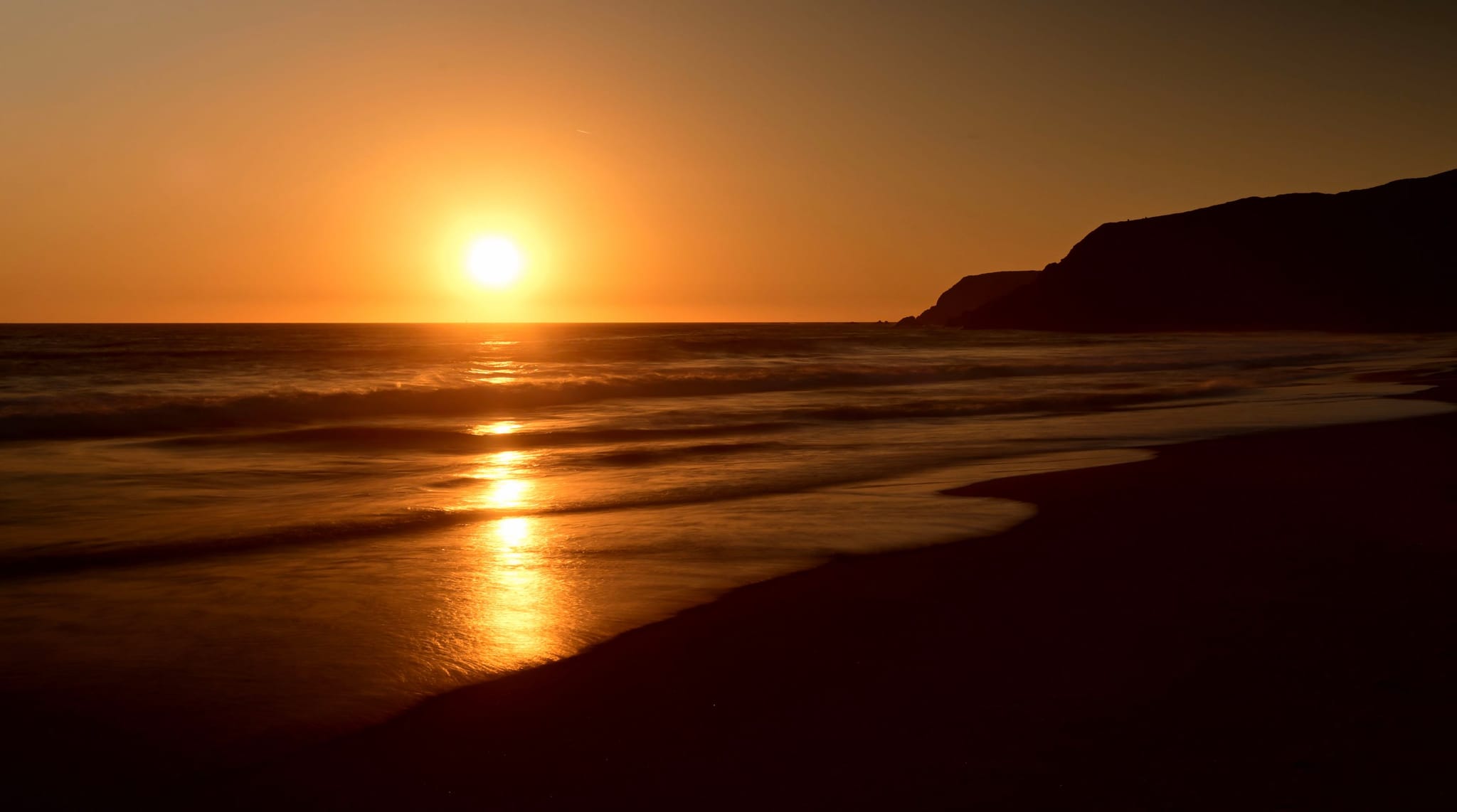 A serene sunset over the ocean with gentle waves lapping against a sandy beach and a silhouette of a distant hill