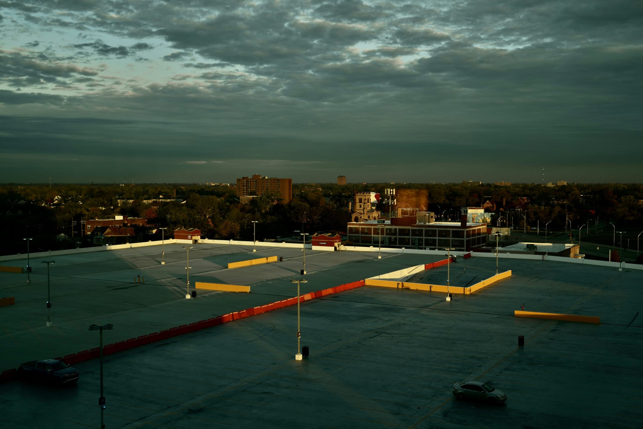 Rooftop parking deck at dusk, yellow barriers glowing in low sunlight, with a cityscape and dramatic clouds in the background